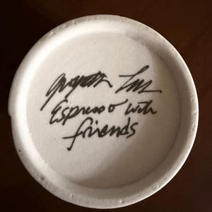 May include: A white ceramic mug base with black handwritten text that reads "Espresso with friends". The mug base has a slightly textured surface and a dark brown background.