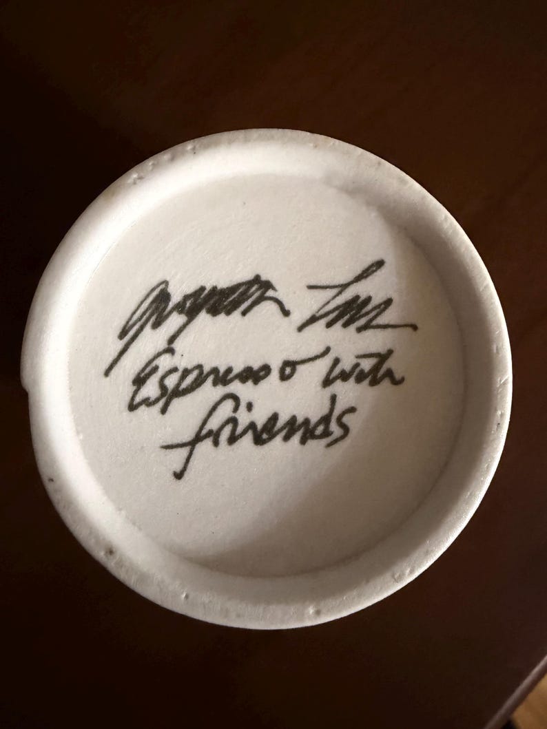 May include: A white ceramic mug base with black handwritten text that reads "Espresso with friends". The mug base has a slightly textured surface and a dark brown background.