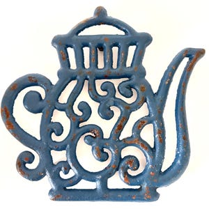 May include: A blue cast iron trivet shaped like a teapot with an openwork scroll design. The metal surface features visible patches of brown rust throughout the blue painted finish, highlighting the texture and age of the decorative kitchen item.