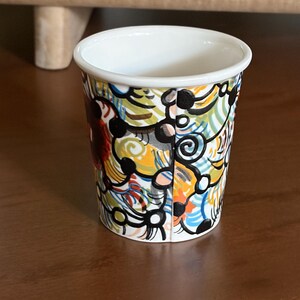 May include: A small, white-rimmed ceramic cup with a colorful abstract design. The cup features a vibrant pattern of swirling lines and shapes in yellow, orange, blue, and black. The cup is set on a dark brown surface.