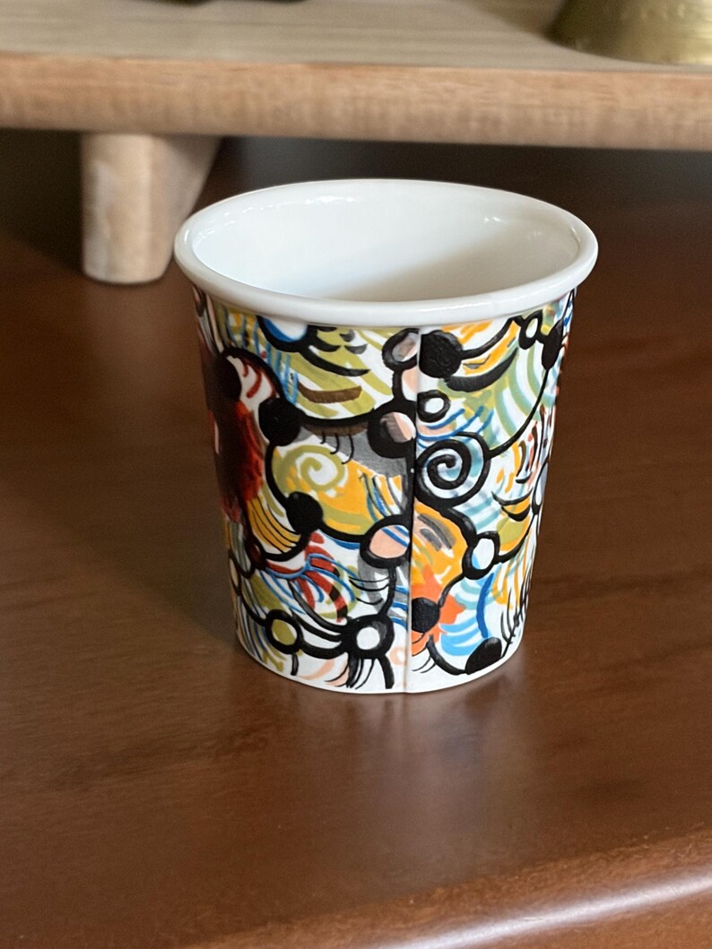 May include: A small, white-rimmed ceramic cup with a colorful abstract design. The cup features a vibrant pattern of swirling lines and shapes in yellow, orange, blue, and black. The cup is set on a dark brown surface.