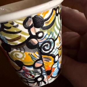 May include: A small, white ceramic cup with a colorful abstract design. The design features black, yellow, orange, blue, and green swirls and shapes. The rim of the cup is white.
