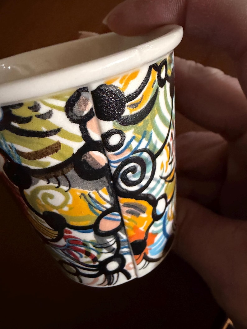 May include: A small, white ceramic cup with a colorful abstract design. The design features black, yellow, orange, blue, and green swirls and shapes. The rim of the cup is white.