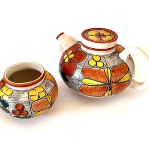 May include: A ceramic teapot with a matching small bowl, both featuring a hand-painted floral pattern in shades of orange, yellow, and dark green against a textured grey background, with a white handle, spout, and interior finish.