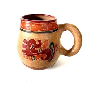 May include: A tan ceramic mug with a rounded body and a thick handle. The rim features a red band with a geometric pattern. A stylized red and black creature design is painted on the front. The surface has a smooth, matte, earthy finish.