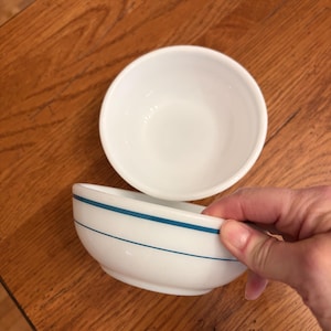 May include: Two white ceramic bowls, one with a blue stripe design. One bowl is upright, and the other is being held. The bowls are on a wooden table. The bowls are likely for serving food.