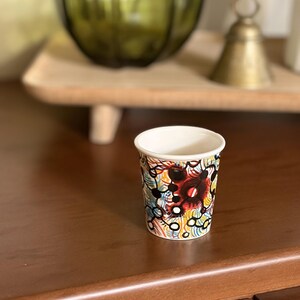 May include: A small, white-rimmed paper cup with a colorful abstract design. The design features red, black, yellow, and blue shapes and lines. The cup is sitting on a dark brown wooden surface.