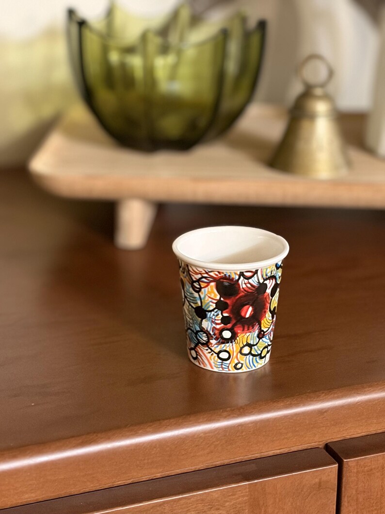 May include: A small, white-rimmed paper cup with a colorful abstract design. The design features red, black, yellow, and blue shapes and lines. The cup is sitting on a dark brown wooden surface.
