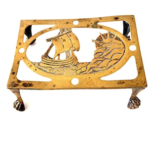 May include: A rectangular brass trivet featuring an openwork design of a sailing ship on the water with a rising sun. The metal stand is supported by four ornate claw-style feet and displays a weathered, aged patina across its surface.