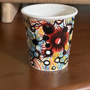 May include: A small, white-rimmed cup with a colorful abstract design. The pattern includes red, black, yellow, blue, and white elements. The cup is cylindrical and sits on a brown surface.