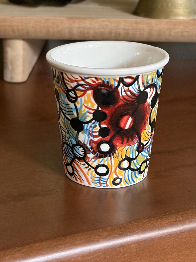 May include: A small, white-rimmed cup with a colorful abstract design. The pattern includes red, black, yellow, blue, and white elements. The cup is cylindrical and sits on a brown surface.