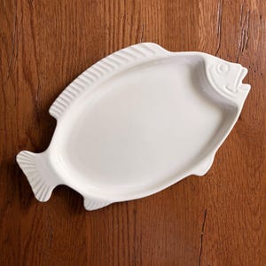 May include: A white ceramic serving platter shaped like a fish. The platter features a smooth, glossy finish with molded details representing the fish's head, fins, and tail. The design is elongated and oval-shaped, providing a flat surface for food.