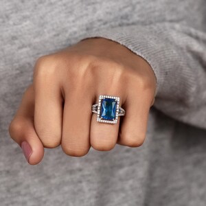 May include: A rectangular blue gemstone ring with a halo of small white stones. The ring has a silver band with additional small white stones on the sides. The ring is set on a hand with a gray background.