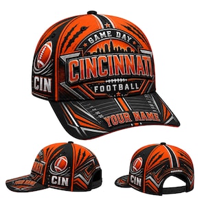 May include: A baseball cap in black and orange featuring the text CINCINNATI FOOTBALL and GAME DAY. The design includes a city skyline, a football graphic, and a field pattern on the brim with the customizable text YOUR NAME in orange letters.