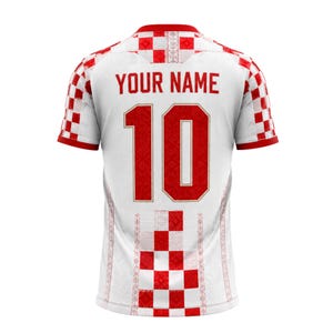 Pu&ograve; includere: Maglia da calcio bianca con dettagli rossi, con il numero 10 in rosso e la scritta "YOUR NAME" sopra. La maglia presenta un motivo a scacchi sulle maniche e sulla parte inferiore della schiena, con un design decorativo sui lati.