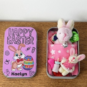 Puede incluir: Una lata de metal rosa con el texto "HAPPY EASTER" y un conejito de dibujos animados sosteniendo un huevo decorado. La lata contiene un conejito de peluche, una almohada y otros juguetes pequeños. La lata está sobre una superficie de madera.