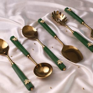 May include: A collection of six gold-coloured serving utensils with green handles. Each handle features a gold-coloured bee. The set includes a ladle, serving spoons, and a pasta server, displayed on a white satin surface.