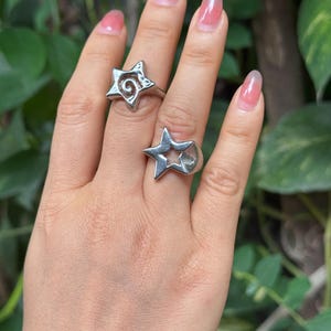 Peut inclure: Deux bagues argentées en forme d'étoile. L'une présente une étoile avec un motif en spirale, l'autre une étoile évidée. Les bagues sont portées sur une main avec du vernis à ongles rose, sur fond de feuillage vert.