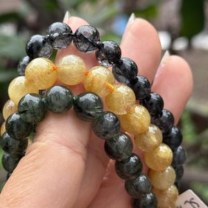 May include: Three beaded bracelets in black, gold, and green. The round beads appear translucent with inclusions. The bracelets are strung on elastic cords, with orange and green accents. The bracelets are held in a hand. The beads are approximately 1 cm in diameter.