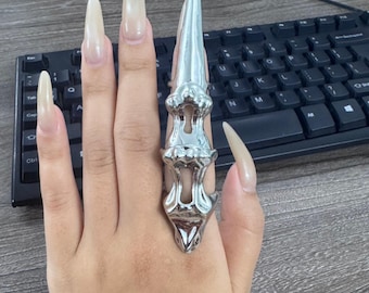 May include: A silver-colored finger ring with a long, pointed design. The ring is worn on a hand with long, light-colored fingernails. The background includes a black computer keyboard.