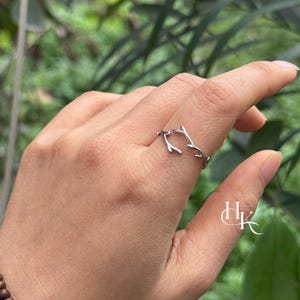May include: A silver-colored ring with a twig or antler design. The ring is open, with the ends of the twig-like design curving around the finger. The ring is worn on a finger, with a blurred green background.