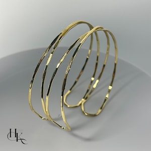 May include: A set of three gold-colored bangles with a hammered texture. The bracelets are stacked together, creating a layered look. The jewelry is displayed on a white surface.