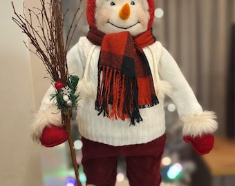 Könnte beinhalten: Eine skurrile Schneemannfigur, gekleidet in einem roten Hut, Schal und Fäustlingen, mit einem weißen Pullover und bordeauxfarbener Hose. Der Schneemann hält einen Zweigbesen und trägt flauschige Stiefel. Ein kleiner Vogelhorst sitzt auf dem Hut.