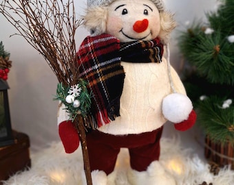 Könnte beinhalten: Eine Schneemannfigur mit grauer Strickmütze, kariertem Schal und roten Fäustlingen. Der Schneemann hat eine Karottennase, einen Reisigbesen und ein kleines Vogelnest auf dem Kopf. Er steht auf einer flauschigen weißen Oberfläche.
