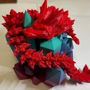 Puede incluir: Escultura impresa en 3D de un dragón rojo con un diseño puntiagudo y texturizado. El dragón está posado sobre una base geométrica con facetas turquesas, azules y moradas. La cabeza del dragón es detallada y su cola se extiende hacia la derecha.