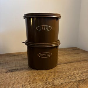 Può includere: Due barattoli marroni impilati. Quello superiore ha la scritta "CAFFE" in un ovale argentato, quello inferiore "ZUCCHERO". Sono su una superficie di legno. Ideali per caffè e zucchero.