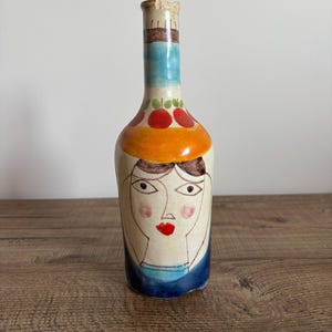 Può includere: Una bottiglia in ceramica dipinta a mano con un design a volto stravagante. La bottiglia presenta una base beige chiaro, un collo blu e una fascia arancione con accenti di frutta rossa. Il viso ha sopracciglia, occhi marroni e un labbro rosso.