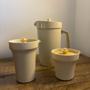 Può includere: Un brocca vintage color crema e due contenitori abbinati con coperchi gialli. La brocca ha un manico e un beccuccio. I contenitori sono cilindrici con una parte superiore leggermente svasata. Il set è esposto su una superficie di legno.