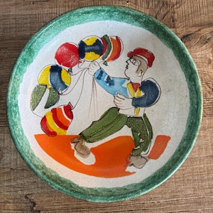 Può includere: Piatto in ceramica dipinto a mano con bordo verde e illustrazione stravagante. L'opera raffigura una figura che tiene in mano palloncini colorati su una base arancione. Il piatto ha uno sfondo bianco con effetto craquelé.