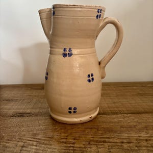 Può includere: Una brocca in ceramica color crema caratterizzata da un motivo dipinto a mano di piccoli fiori blu a quattro petali disposti in file attorno al corpo. Il recipiente presenta un manico curvo sul lato destro e un beccuccio pronunciato.
