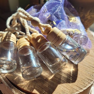 May include: Four small, clear glass bottles with wooden caps and hanging cords, arranged on a wooden surface. A sheer purple bag with gold accents is in the background. The bottles appear to contain a clear liquid.
