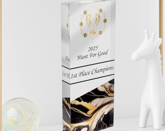 Puede incluir: Un trofeo rectangular transparente con detalles dorados y el texto "2025 Hunt For Good 1st Place Champions". El trofeo tiene un diseño de mármol negro y dorado en la base y un diseño circular dorado en la parte superior.