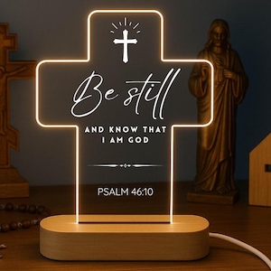 May include: A cross-shaped LED night light with the words "Be still and know that I am God" and Psalm 46:10. The light has a wooden base and a white cord. Other religious items are in the background.