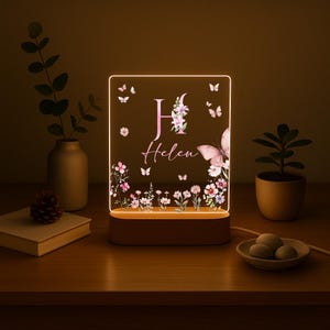May include: A personalized LED night light with a floral and butterfly design. The light features the name "Helen" and the initial "H" in pink, surrounded by flowers and butterflies. The base is brown, and the light emits a warm glow.
