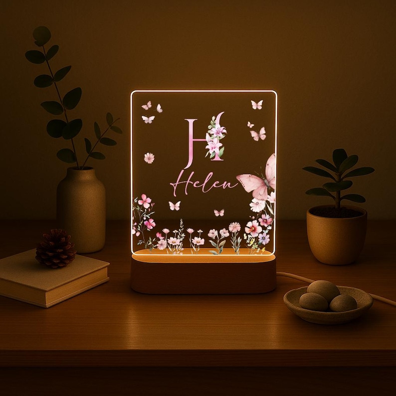 May include: A personalized LED night light with a floral and butterfly design. The light features the name "Helen" and the initial "H" in pink, surrounded by flowers and butterflies. The base is brown, and the light emits a warm glow.