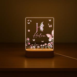 May include: A personalized night light with a floral and butterfly design. The acrylic panel features the name "Helen" and the letter "H" in pink, surrounded by flowers and butterflies. The light sits on a wooden base and is connected to a power cord.