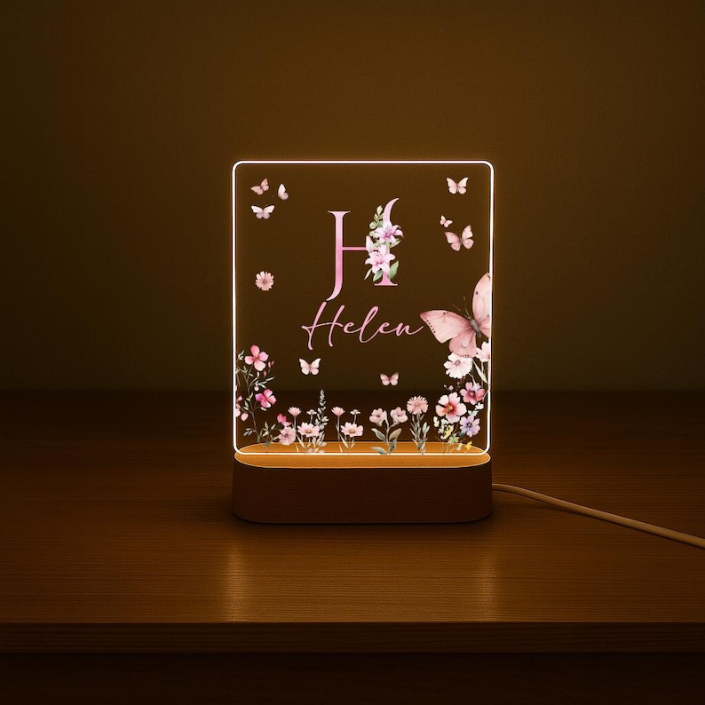 May include: A personalized night light with a floral and butterfly design. The acrylic panel features the name "Helen" and the letter "H" in pink, surrounded by flowers and butterflies. The light sits on a wooden base and is connected to a power cord.