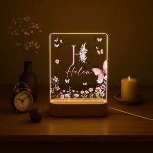 May include: A personalized night light with a wooden base. The acrylic panel features a floral design, butterflies, and the name "Helen" with the letter "H". The light is illuminated with a warm glow, creating a cozy ambiance.