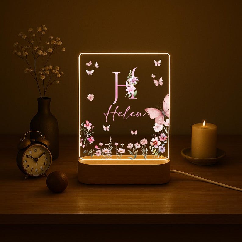 May include: A personalized night light with a wooden base. The acrylic panel features a floral design, butterflies, and the name "Helen" with the letter "H". The light is illuminated with a warm glow, creating a cozy ambiance.