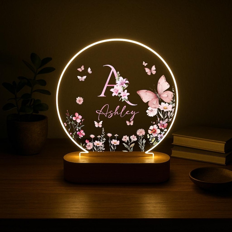 May include: A personalized night light with a circular design. The light features the name "Ashley" and the letter "A" in pink, surrounded by floral and butterfly illustrations. The base is a dark wood color, and the light emits a warm glow.