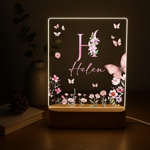 May include: A personalized night light with a wooden base. The acrylic panel features a floral and butterfly design with the name "Helen" and the letter "H" in pink. The light emits a soft glow, perfect for a bedroom.