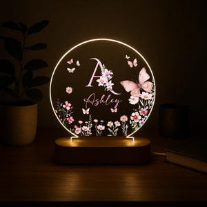 May include: A personalized night light with a circular design. The light features a pink butterfly and floral design, with the name "Ashley" and the initial "A" in pink. The light sits on a wooden base.