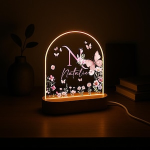 May include: A personalized night light with a wooden base and an arched acrylic panel. The panel features a pink floral design, butterflies, the letter "N", and the name "Natalie" in a script font. The light emits a warm glow.