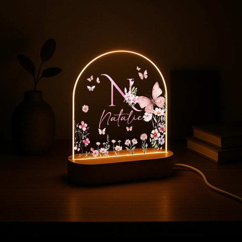 May include: A personalized night light with a wooden base and an arched acrylic panel. The panel features a pink floral design, butterflies, the letter "N", and the name "Natalie" in a script font. The light emits a warm glow.