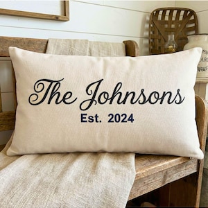 May include: Cream-colored rectangular pillow with the words "The Johnsons" in black script and "Est. 2024" in navy blue. The pillow is resting on a wooden bench with a neutral-toned fabric.