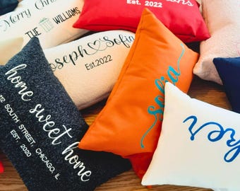 May include: A collection of personalized throw pillows in various colors, including red, orange, white, and navy. The pillows feature embroidered text such as "Merry Christmas," "The Browns Est. 2022," "Joseph & Sofia Est. 2022," and "home sweet home 112 South Street, Chicago, IL EST. 2020."